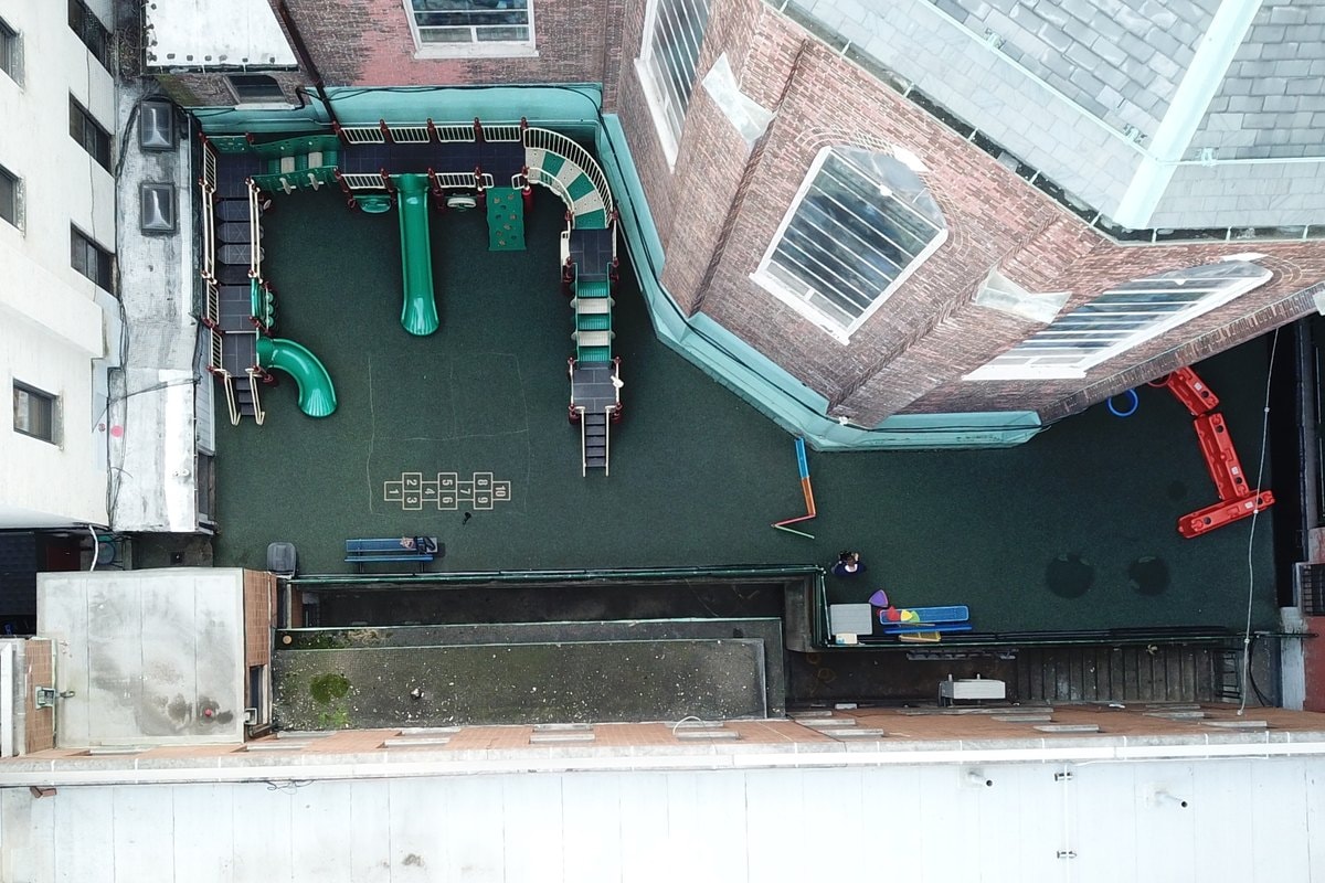 Straight-down drone shot of the old green-surface courtyard playground with existing teal and red equipment.