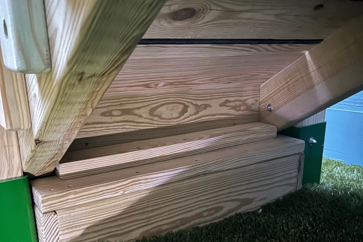 Close-up under the deck — green steel post brackets, wood stringers, blue siding visible at right, artificial turf below.