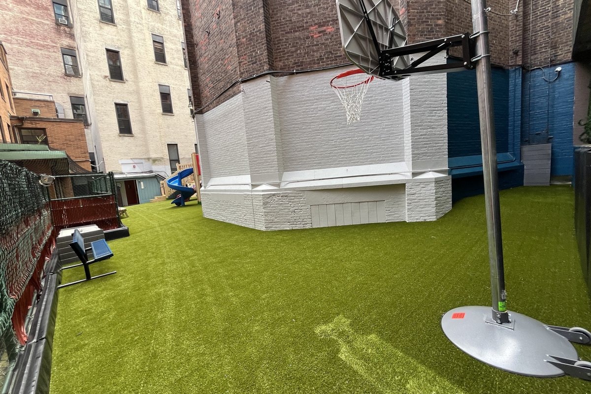 Wide shot of the finished courtyard — basketball hoop, artificial turf, playground visible in background.