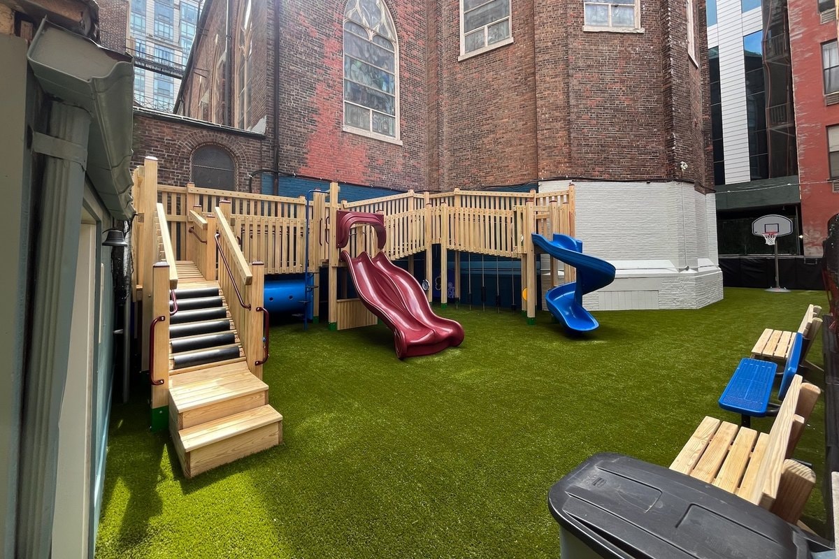 Slightly elevated angle — full courtyard with entry stairs at left, both slides, basketball hoop at right.