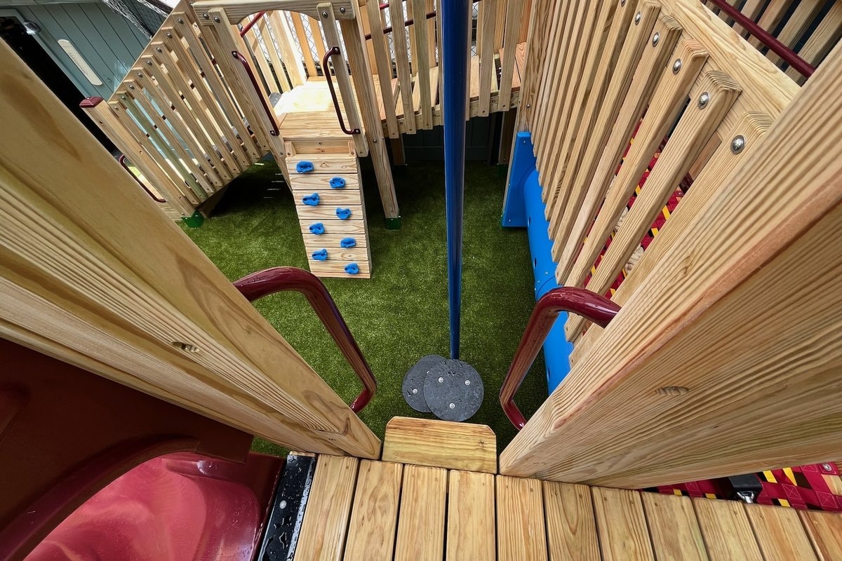 Looking straight down from the upper deck — blue climbing wall with holds at left, red spiral slide entry at bottom, blue tunnel at right.