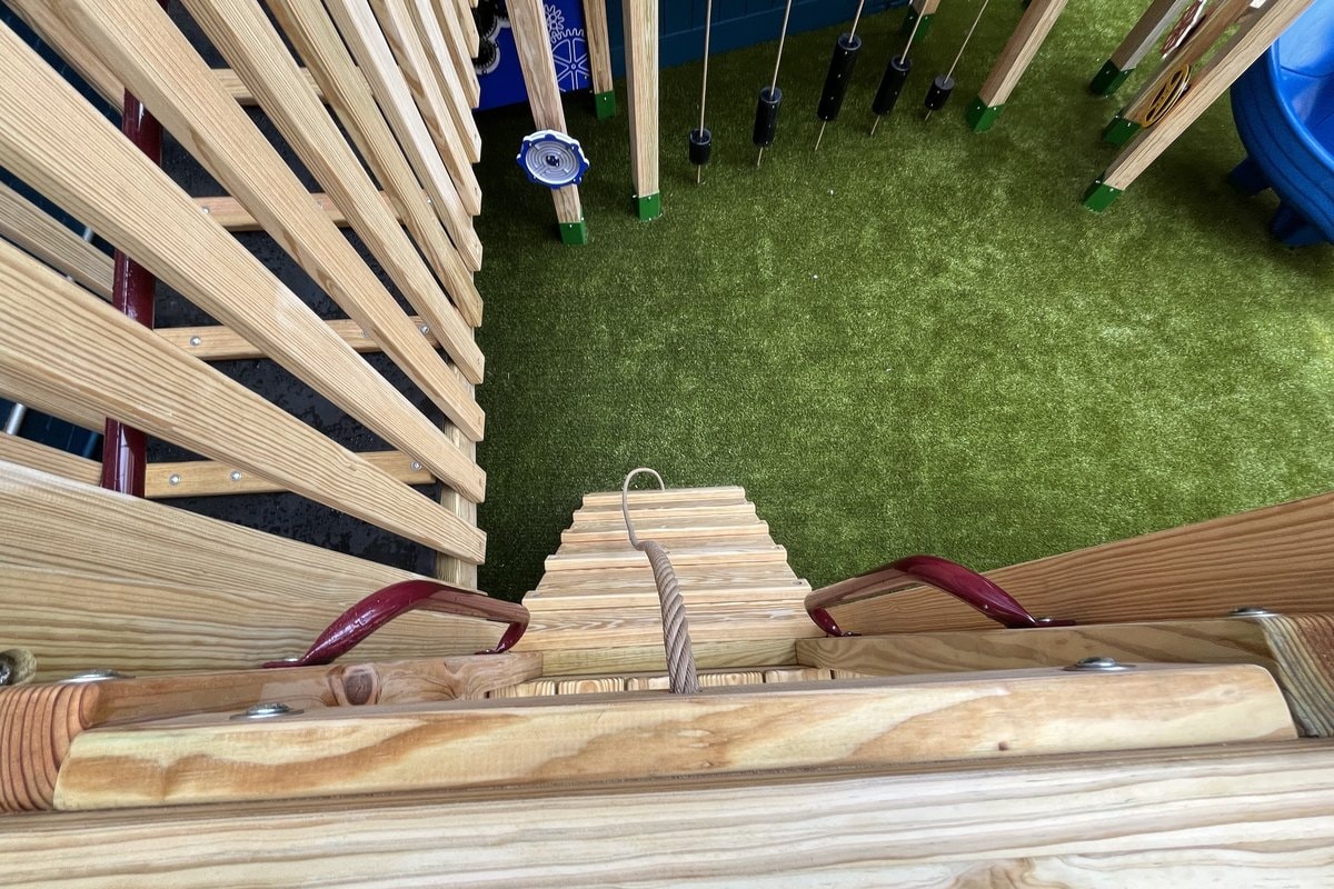 Looking straight down from upper deck — wooden rope ladder descending to turf, play panels with spinners visible in background.