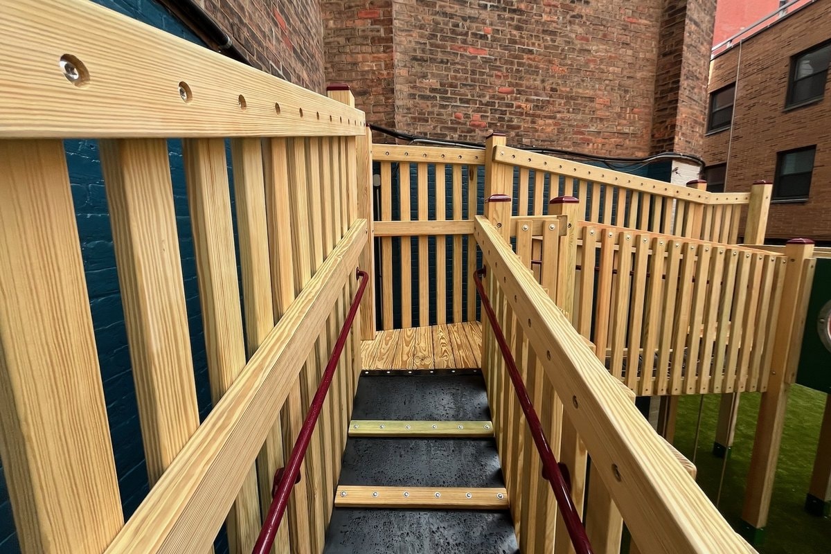 Ground-level view of the main entry staircase — natural wood treads, burgundy steel handrails, brick building backdrop.