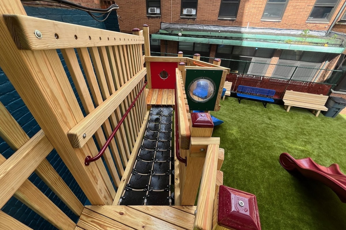 From upper deck looking down — rope bridge leading to red and green porthole panels, blue and wooden benches visible in background.