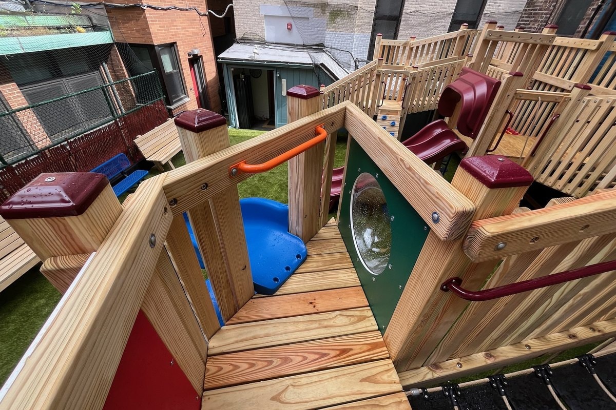 From upper deck looking down at lower deck — wood plank walkway, green panel with porthole, blue rocker at left, red slide entry at right.