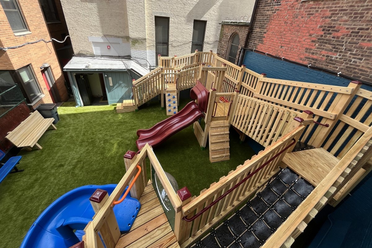 Wide elevated shot of the full courtyard — burgundy slide center, blue spiral slide right, multi-level wood deck, artificial turf, brick surrounds.