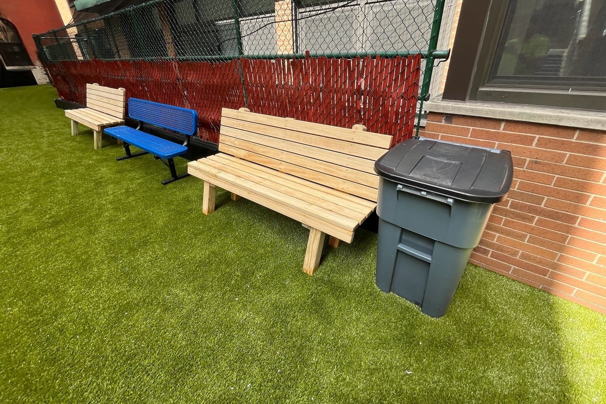 Ground-level shot of the bench area — two natural wood benches and one blue metal bench against the fence, artificial turf, brick building at right.