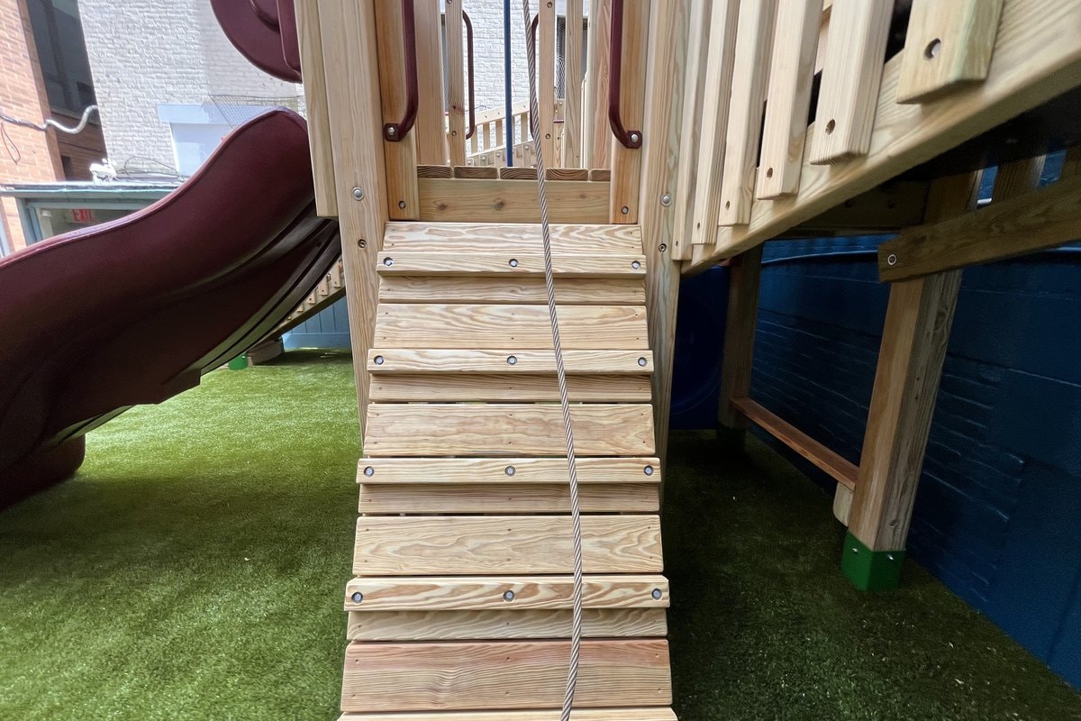 Ground-level view of the rope-assisted ramp leading up to the deck — natural wood treads with rope guide rail, burgundy slide visible at left.