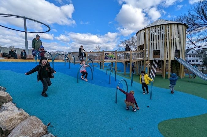 Children of different ages running and playing across a playground in front of a wooden tower.